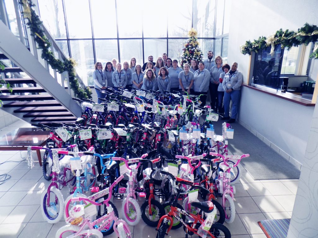 A group of Trans-Overseas employees stands in front of a  large plate glass windows and behind 43 children's bikes for the DIA Toys for Tots Charity event supported by Trans-Overseas Corporation. A Staircase ascends on the left side of the frame towards the camera while the bikes are arranged in 4 rows of 10 with 3 bikes in the immediate foreground. The receptionist's window is on the right of the frame.